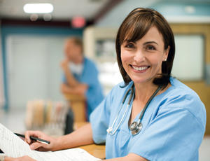 Femme médecin portant un uniforme bleu et un stéthoscope autour du cou. Elle tient un stylo et du papier. Elle sourit.