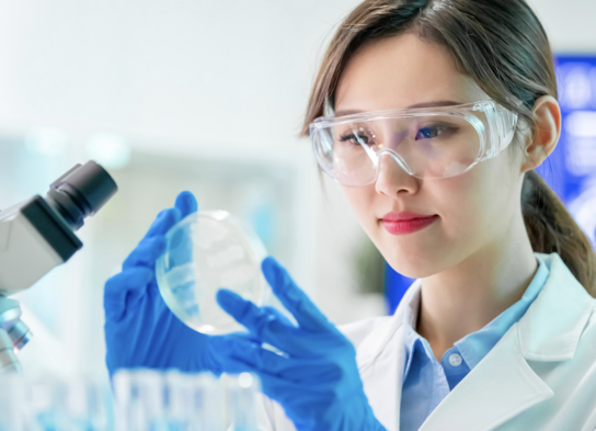 Woman wearing laboratory gloves and security glasses manipulating laboratory flask with pink liquid inside.