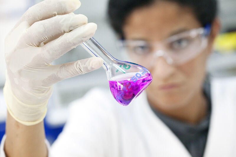 Woman wearing laboratory gloves and security glasses manipulating laboratory flask with pink liquid inside.